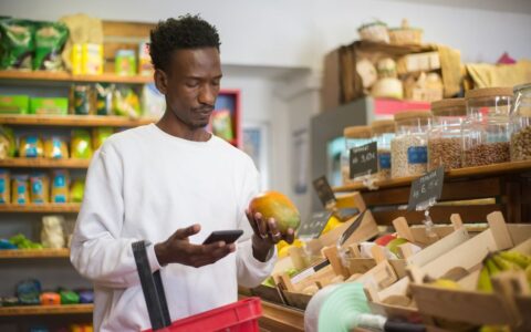 A man examines a mango while shopping with a smartphone in a grocery store.