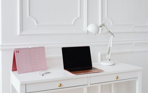 Simple desk setup with laptop, calendar, and lamp in a bright, minimalist office setting.
