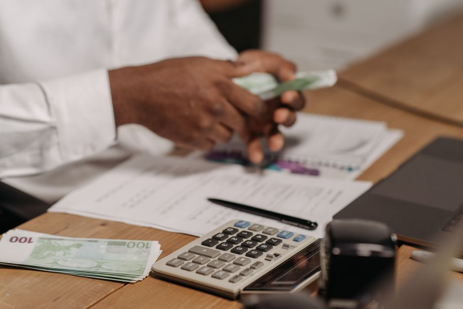 Hands counting euro bills on a wooden desk with calculator, financial documents, and laptop nearby.