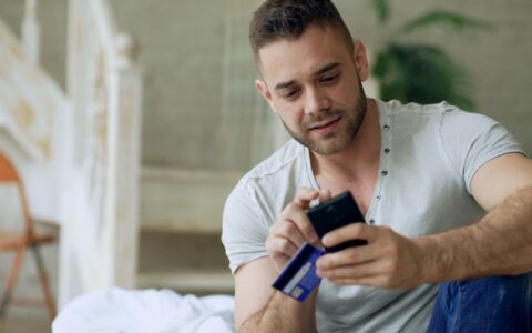 Young man using smartphone and credit card for online shopping indoors.